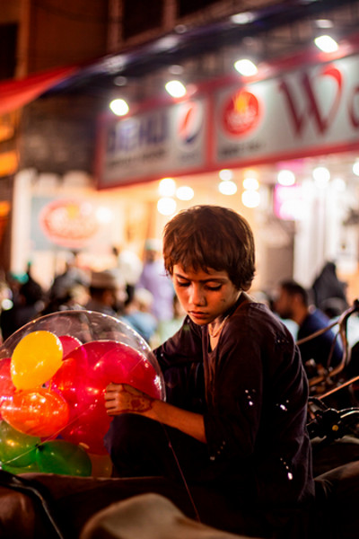  Taken in Lahore, Pakistan on December 15. This photograph depicts an emotional intercultural moment, with a young boy in a bustling street scene in Lahore, Pakistan. The youngster, adorned in customary clothing, is carefully inspecting a cluster of colourful balloons. The child's serious countenance juxtaposed with the vibrant balloons represents a combination of innocence and the hard truths of a life dedicated to street work. The setting includes a combination of Eastern and Western civilizations, demonstrated by the bilingual signs and the fusion of traditional and contemporary components. This image depicts the meeting point of worldwide cultures and personal narratives within a broader societal framework. The boy's image is an intercultural moment due to its cultural attire, global symbols, urban melting pot, economic diversity, the blend of tradition and modernity, and linguistic interculturalism. The boy's traditional attire represents his cultural heritage, while the balloons symbolize celebration. The setting is an urban environment where multiple cultures coexist and interact, reflecting the intersection of local life with global influences. The boy's solemn demeanour and selling balloons reflect global issues like child labour and economic disparities.